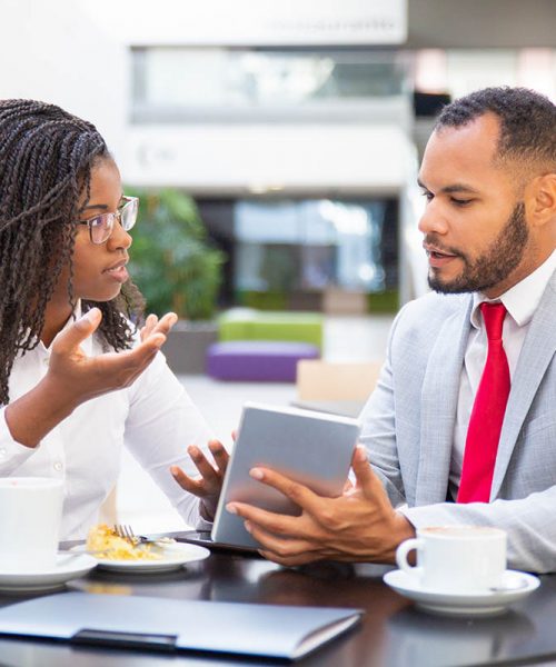 Businessman showing project presentation to female colleague. Diverse business man and woman sitting in cafe, using tablet together and talking. Digital communication concept